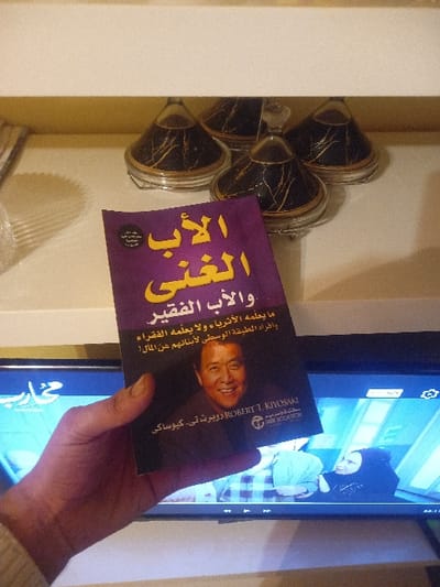The image displays a hand holding a purple book titled "Rich Dad Poor Dad" in Arabic. The book cover features a photo of Robert T. Kiyosaki. The text on the cover reads, "What the Rich Teach Their Kids About Money - That the Poor and Middle Class Do Not!" The backdrop includes a television screen showing an image of a woman in a hijab, along with a shelf containing several decorative items.