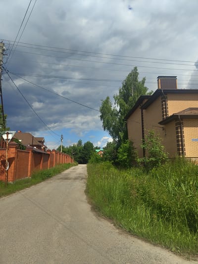 The image captures a view of a dirt road curving towards the right in the village of Tsibino, Russia. On the left, a long, red brick wall with decorative caps lines the road, separating it from a residential property with a brown-roofed house visible behind it. Overhead, power lines crisscross the sky, supported by a utility pole on the left. The right side of the road is bordered by tall, lush green grass and foliage. Further down the road, more trees and houses are visible, suggesting a suburban or rural residential area. The sky is partly cloudy, with dark storm clouds gathering in the upper left quadrant, contrasting with patches of blue sky. The scene appears to be a typical summer day, possibly before or after a rain shower.