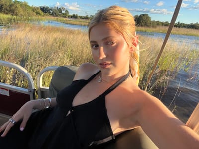 A woman is sitting in what appears to be a boat, possibly a pontoon boat, on a body of water. She is wearing a black dress and jewelry, including a necklace, bracelets, and earrings. The background shows a landscape with tall grasses and trees. The location is likely near the city of Victoria Falls, Zimbabwe, given the presence of such a lush landscape, suggesting the Zambezi River area or a nearby wetland. The lighting suggests either early morning or late afternoon.