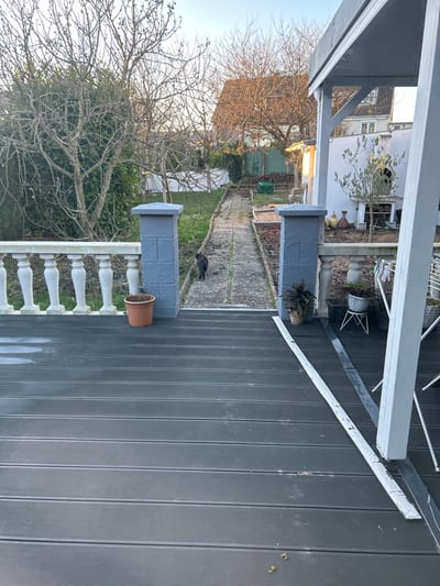 The scene depicts a residential backyard or garden in Saint-Quentin, France, viewed from a dark grey composite deck. The deck features a white balustrade with ornate spindles on the left, flanked by two grey block pillars. A terracotta pot rests on the deck near the left pillar. A long, light-colored strip and a darker metallic strip lie horizontally on the deck surface towards the right. A grey support pillar for an overhead structure stands on the far right, near which a potted plant with dark leaves and a white metal plant stand with additional potted plants are visible. A white clothes drying rack is partially obscured on the far right.
Beyond the deck, a concrete or stone slab path, partially covered by moss, leads through the garden. A black cat is walking away from the viewer down this path. The garden features sparse vegetation and bare deciduous trees, indicating a late autumn or winter setting. A large evergreen shrub is prominent on the left. On the right side of the garden, against a light-colored wall, an outdoor oven or fireplace with stacked firewood is present, accompanied by several colorful ceramic pots and a small, green tree. In the background, other residential structures with tiled roofs are visible under a clear, pale blue sky. No people are present.