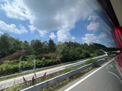 The image depicts a view from inside a vehicle, likely a bus or car, traveling on a highway. The highway has two lanes and is bordered by a metal guardrail on both sides. Lush green trees and vegetation line the left side of the highway, with a gentle slope leading up to a dense forest. The sky is partly cloudy with scattered white clouds against a bright blue backdrop. On the right side of the image, the reflection of the vehicle's interior is visible on the window, including a red curtain. The road appears to be an elevated section of the highway, with a curved overpass visible in the distance. The scene is set in a natural, rural environment, suggesting a journey through the countryside.