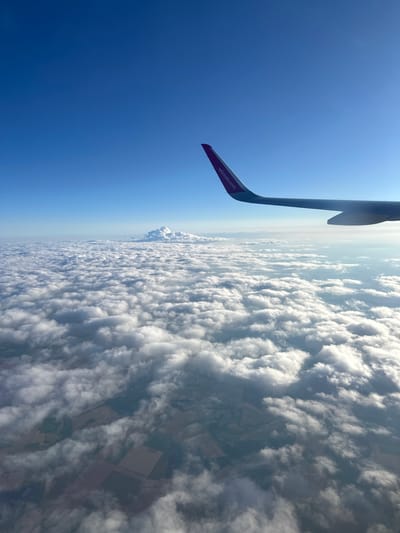 An aerial view from an airplane window shows a vast expanse of white clouds stretching across the horizon. The wing of the aircraft, with "wizzair.com" printed on it, extends into the upper right corner of the frame. Below the clouds, hints of agricultural fields and land are visible, suggesting a rural or semi-rural landscape. In the distance, a towering mountain range, likely the Balkan Mountains, is partially obscured by a large, cauliflower-like cloud formation. The sky is a clear, deep blue. The image captures the scene above the city of Vekilski, Bulgaria.