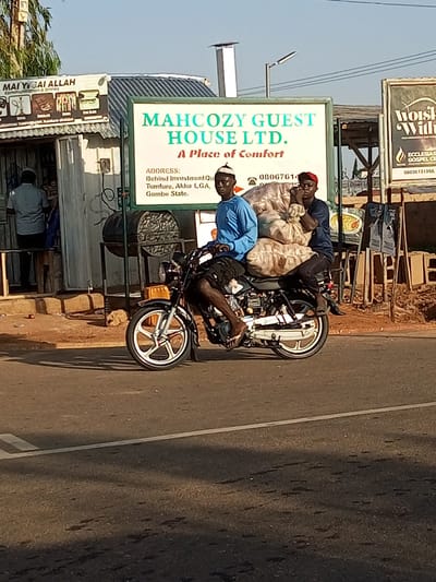 Two men are riding on a motorcycle, carrying large sacks of goods. The man in front is wearing a blue t-shirt and a white cap. The man behind him is wearing a dark blue t-shirt and a red cap. They are on a paved road in front of a building with a sign that reads "MAHCOZY GUEST HOUSE LTD. A Place of Comfort". There is another sign to the right that reads "Woisi Wilh" and "ECCLESIASI GOSPEL CH". The scene is likely in Wuro Dole, Nigeria, as indicated by the context. The time of day appears to be late afternoon or early evening due to the long shadows. The environment is an urban or semi-urban setting, with buildings and electrical wires visible.