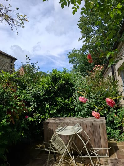 A low-angle, eye-level shot captures a charming garden scene in Middleton Tyas, United Kingdom. The foreground is dominated by a light-colored, foldable metal table and two chairs, casting long shadows on the stone patio. Behind the seating area, a rustic wooden structure, possibly a planter box or storage unit, stands prominently.

The background is lush with greenery, featuring dense bushes and trees. Several pink roses in bloom are scattered throughout the foliage, adding pops of color. In the upper left, the corner of a stone building with a tiled roof is visible. Towards the center background, a brick chimney rises above the greenery. The sky above is a mix of blue with scattered white clouds. The overall impression is one of a serene, sunlit garden.