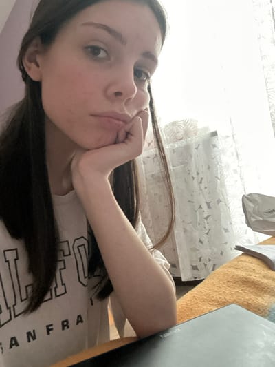 An indoor, eye-level photograph depicts a young adult female in the foreground, positioned slightly to the left of center, looking directly at the camera. Her dark, straight hair falls over her shoulders. She wears a white t-shirt with partial dark text reading "CALIFORNIA SAN FRANCISCO". Her right hand supports her chin, and she maintains a neutral expression. In the bottom right foreground, a dark digital tablet, possibly an X-Pen brand, rests on a surface covered by an orange-yellow textured fabric. Behind the female, a brightly lit window is visible, covered by translucent white curtains featuring a repeating floral or geometric pattern. To the right of the female, near the window, a crumpled white object and some papers are discernible on the same fabric-covered surface. The wall to the left of the female is a solid light purple hue. The scene is consistent with a residential interior in the city of Волховец, Russia.