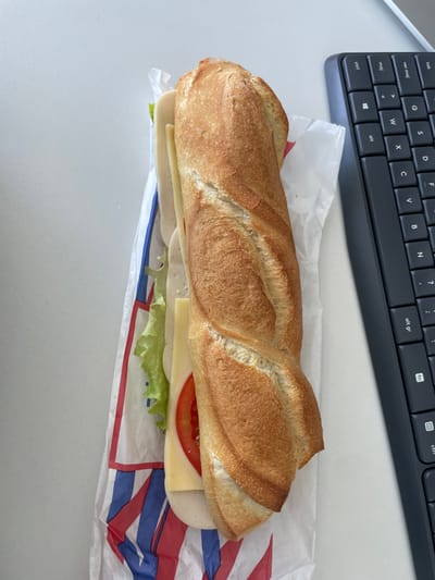 An overhead image depicts a baguette sandwich positioned on a white, unadorned surface, likely a desk, within Montigny-le-Bretonneux, France. The sandwich lies horizontally, slightly angled towards the top right. It consists of a golden-brown, crusty baguette split lengthwise, revealing visible fillings: green ruffled lettuce, several slices of light-colored deli meat (such as turkey or ham), a slice of light yellow cheese, and a red tomato slice. The sandwich is partially encased in white paper packaging, which displays a red and blue striped pattern on its left side. To the immediate right of the sandwich, a portion of a dark-colored computer keyboard is visible, featuring keys such as 'm', 'l', 'ctrl', 'alt', 'f11', and 'f12', consistent with an office or personal computing environment and likely an AZERTY layout. No individuals or active interactions are visible.