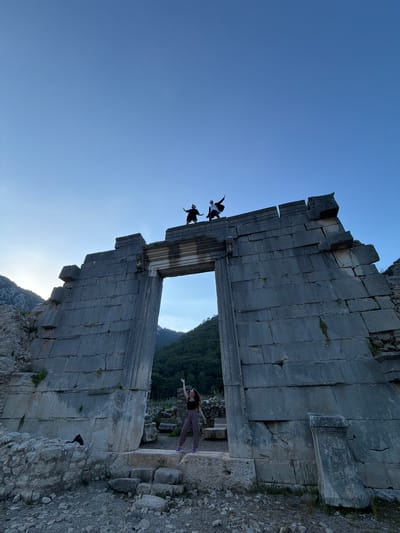 An outdoor, wide-angle shot captures an ancient stone structure against a clear blue sky, featuring three individuals engaging with the ruins, likely near Kumluca, Turkey. The image is rotated 90 degrees counter-clockwise, making the structure appear to the right and left of the frame when described from an upright perspective.
The central subject is a large, grey stone wall, likely part of a historical building or gateway, with a prominent archway or doorway. The stonework is rough-hewn and appears ancient, showing signs of wear and crack.
Two people are silhouetted against the sky, standing on the very top edge of the stone structure to the left of the archway. They are both positioned with their arms spread wide, giving the impression of balancing or "flying" as they pose.
A third person is visible inside the archway on the right side, seen from behind and facing away from the camera. This individual also has their arms outstretched, creating a similar pose to the figures on top. They are wearing dark clothing and light-colored pants.
Through the archway and beyond the structure, a landscape of green, tree-covered mountains or hills is visible, extending into the distance under the bright sky. A low sun, possibly setting, casts a warm light near the base of the archway, creating a subtle glow. The ground surrounding the ruins is rocky and uneven. The overall atmosphere suggests a clear, pleasant day, likely late afternoon or early evening, at a historical site. There is no visible text in the image.