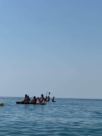 In the bright, clear waters off Finale Ligure, Italy, several people are enjoying a day on the sea. A yellow buoyant marker is visible on the left side of the frame, with choppy blue water extending towards the horizon. In the mid-ground, a group of people are gathered on a flat, yellow raft. Some are seated, while others are in various relaxed postures. Two individuals are seen standing on paddleboards further out in the water, one with a paddle raised. The sky above is a clear, light blue, indicating fair weather conditions. The scene suggests a leisurely recreational activity on a calm sea.