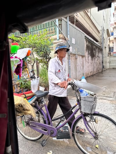 A person, possibly a vendor, is resting on a purple bicycle in a narrow alley. They are wearing a white long-sleeved shirt, dark pants, and a cap, and appear to be asleep or taking a break. The bicycle is equipped with a basket in the front containing two water bottles and a large, flat piece of plastic or metal, possibly a shield or awning. Attached to the rear of the bicycle are various cleaning supplies, including brooms with different colored bristles and what appears to be a white mop. The scene is set in an urban environment, likely in Khan Chamkar Mon, Cambodia, judging by the architecture of the buildings lining the alley. The buildings are multi-story with light-colored facades and visible air conditioning units. The ground is paved with concrete, and there are some debris and loose papers scattered around. Green foliage from a nearby tree or plant is visible, adding a touch of nature to the otherwise utilitarian setting. The lighting suggests it could be daytime, but the overall atmosphere feels subdued, hinting at a break from activity. The presence of cleaning tools and the way the person is positioned on the bicycle might indicate they are a street vendor or a cleaner who uses the bicycle for transport and storage of their goods. There is no visible text in the image.