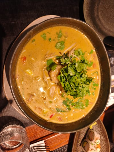 A close-up, overhead shot captures a bowl of Thai yellow curry soup with shrimp, bean sprouts, and cilantro. The soup is served in a dark green, speckled bowl, which sits on a light brown charger plate. To the left, a clear glass with a ribbed design is partially visible, along with a fork. In the lower right corner, a small plate holds a couple of sushi rolls. The scene is set on a wooden table with a dark, textured surface. The lighting suggests an indoor setting, possibly a restaurant. The image was taken in Bnei Dror, Israel.
