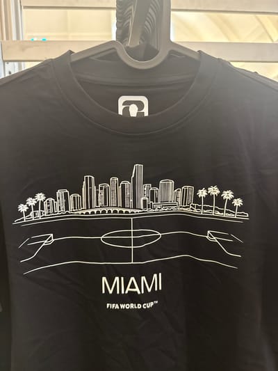A black, long-sleeved t-shirt is displayed on a dark grey plastic clothes hanger, viewed from a slightly elevated frontal perspective within an indoor setting in Sunrise, United States of America. The t-shirt's ribbed crew neck shows a white brand tag with a stylized dark grey logo, resembling an abstract 'P' or '6'. The garment features a prominent white outline graphic depicting a cityscape, specifically Miami, with multiple tall buildings, a bridge, and groups of palm trees flanking the skyline. Below the cityscape, a stylized football (soccer) pitch outline is present, showing the center circle, halfway line, and partial goal areas. Beneath this graphic, two lines of white text are centered: "MIAMI" in large uppercase letters, followed by "FIFA WORLD CUP™" in smaller uppercase text. The t-shirt fabric shows minor creasing. The background is out of focus, revealing horizontal light-colored elements consistent with blinds or shelving, with indistinct light sources and two small blue illuminated points in the upper left quadrant.