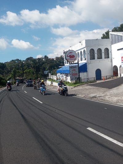 The image depicts a daytime scene on a paved road in Ban Phonhxavanh, Laos, under a clear blue sky with scattered white cumulus clouds. The sun casts long shadows from left to right across the asphalt road, which features a solid white line on its right edge. Multiple motorcycles and one black car are observed moving on the road from left to right. In the foreground, a person wearing a white helmet rides a dark motorcycle, followed by another dark motorcycle. Further right, a black car is observed. A cluster of several motorcycles follows the car, with at least one rider wearing a yellow helmet. In the mid-ground, a person in a yellow helmet operates a light blue motorcycle. On the far right foreground, two individuals are on a dark grey scooter-style motorcycle; the driver wears a dark helmet, and a passenger is seated behind.

On the right side of the road, a multi-story white building with classical architectural elements, including multiple arched windows and a dark blue arched doorway, is prominent. A large circular sign for "D'Colonel," featuring a cartoon chef holding a chicken, is affixed to the building's facade. Below this, a blue canopy extends, under which a rectangular sign with text including "DC... PROJECT" and "TODAY" is visible. Green foliage lines the paved area in front of the building, which includes grey interlocking bricks leading to a darker paved surface. An orange traffic cone is placed on the paved area near a blue door on the building. To the right of this structure, another section of a white building with a more angular, possibly corrugated, roof design is partially visible. Lush green trees and vegetation cover hills in the background, behind the buildings.