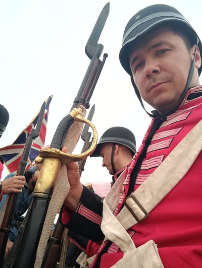 The image displays a group of individuals in historical military attire, positioned outdoors in Kottalapalli, India. In the foreground, a male subject wears a black helmet with a chin strap, a red military-style jacket with black trim and light-colored horizontal chest stripes, and a beige cross-belt. He looks directly at the viewer. His right hand grasps the golden hilt of a sabre or sword, which is positioned in front of a vertically held firearm fitted with a bayonet. Behind and to the left of the foreground subject, a second male, also in a black helmet and similar uniform, is partially visible, facing left. Further left, a third, largely obscured individual wearing a blue denim shirt holds a pole from which a Union Jack flag is displayed. Several other firearms with attached bayonets are visible vertically throughout the composition, held by indistinct figures. The background consists of a uniform, light overcast sky.