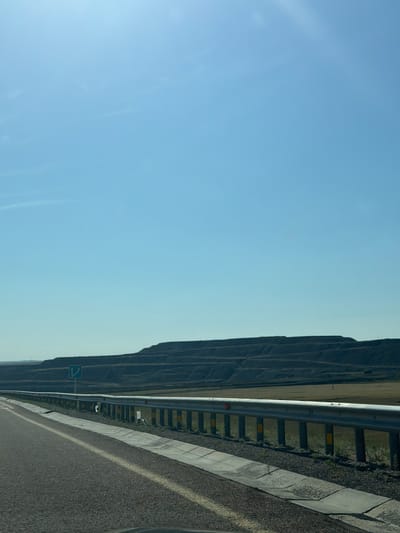 The image captures a view from a moving vehicle on a highway near Ereymentaw, Kazakhstan. The foreground shows the asphalt road with a yellow line marking the lane. A metal guardrail runs along the side of the road, separating it from a concrete drainage channel and then a grassy embankment. In the middle ground, a large, terraced hillside is visible, likely a mine or quarry, with distinct layers of earth. The background consists of a vast, clear blue sky with subtle wisps of clouds. A road sign with blue arrows indicating a lane split is also visible to the left of the highway. The overall scene is one of a rural or semi-industrial landscape under a bright sky.