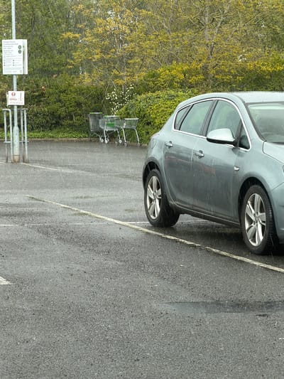 The image shows a gray car parked in a parking lot in Pity Me, United Kingdom. The parking lot is lined with a row of trees and bushes. There are several shopping carts stacked next to the parking lot. There is a sign that reads "Arnison Centre Customer Car Park Maximum stay 6am-10pm Maximum stay 10pm-6am." The parking lot appears wet, and it may be raining.