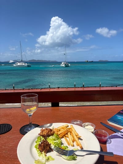 The image depicts an outdoor dining scene in Spanish Town, British Virgin Islands, overlooking a tropical bay. The foreground features a reddish-brown wooden table with a partially consumed meal on a white oval plate. The meal consists of several fried, shredded fritters on lettuce and a side of French fries. A silver fork rests on the plate. To the right of the plate are three small clear plastic condiment cups containing red ketchup, white mayonnaise, and an unidentifiable third substance. A silver knife, a folded purple cloth napkin, and a dark-covered book are also on the table. To the left, a clear stemmed glass contains a beverage and a lemon wedge, resting on a black textured coaster.
A wooden railing adorned with a string of white miniature decorative lights separates the table from the water. Beyond the railing, a sandy shoreline is visible before the clear turquoise ocean. The background features a vibrant blue sky with large white cumulus clouds. Several boats are present in the water: two prominent white catamaran sailboats, a large white motor yacht further left, a smaller personal watercraft near the center, and a larger, darker vessel in the distant right. Distant islands or landmasses are visible on the horizon. The scene is illuminated by bright daylight. No individuals are visible.