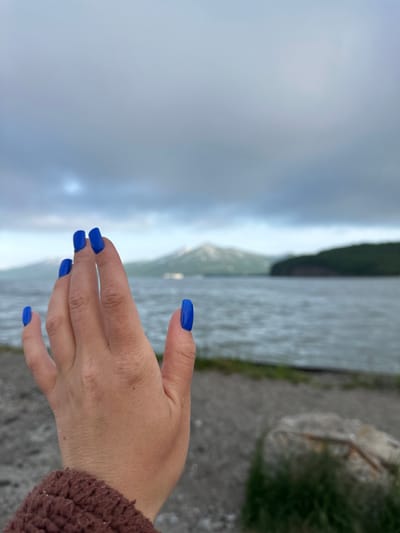 A hand with bright blue polished fingernails is held up in the foreground, obscuring part of the view. The hand is oriented towards the viewer, with fingers slightly spread. In the background, a body of water, likely a bay or strait, is visible, with choppy waves indicating wind. Across the water, a range of hills and mountains are partially obscured by clouds and mist. The landscape appears rugged and natural. The sky is overcast and a dull gray, suggesting cloudy weather. The foreground shows a gravelly or sandy shore with some sparse green vegetation and a large rock. The image was taken in Vilyuchinsk, Russia, a city known for its proximity to volcanoes and its naval base. The overall scene evokes a sense of raw, natural beauty under a dramatic sky.