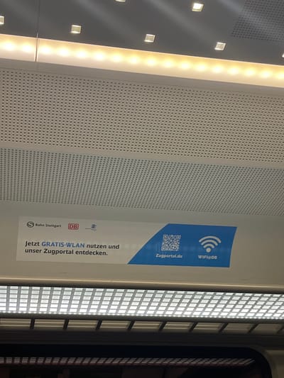 The image displays a forensic view of the interior ceiling and upper wall of a modern train carriage, located within Echterdingen, Germany. The ceiling features light grey, perforated panels designed for acoustics or ventilation. Overhead illumination is provided by two distinct lighting fixtures: an upper linear strip of warm-toned LED lights interspersed with square recessed spotlights, and a lower linear fixture covered by a grid diffuser, emitting white light. Affixed to the upper wall below the lighting is a white and blue rectangular adhesive sign. This sign prominently displays the logos for "S Bahn Stuttgart" and "DB," along with a third, smaller, stylized logo. The German text on the sign reads, "Jetzt GRATIS-WLAN nutzen und unser Zugportal entdecken," which translates to "Now use FREE Wi-Fi and discover our train portal." The right side of the sign, in blue, features a QR code, the web address "Zugportal.de," and a Wi-Fi symbol accompanied by "WIFI@DB." No individuals, active interactions, or specific seating arrangements are visible within the captured frame.