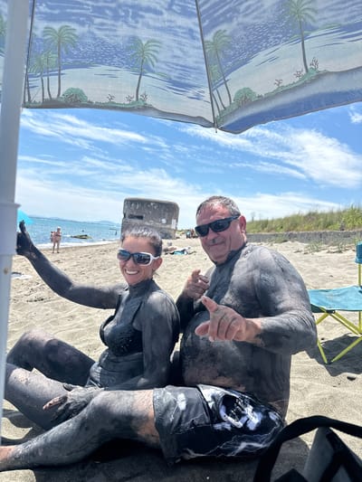 In a sun-drenched scene on a sandy beach, two individuals, a man and a woman, are covered head-to-toe in dark mud. They appear to be participating in a mud bath, a popular activity in Aheloy, Bulgaria. Both are wearing sunglasses, and the woman has a bright smile, suggesting they are enjoying the experience. The man is leaning towards the camera, his hand extended, as if interacting with it or gesturing. The woman is positioned slightly lower, her arm bent at the elbow.

The background reveals a tranquil sea under a bright blue sky dotted with wispy white clouds, indicative of pleasant weather. The beach itself is sandy, with a few people in the distance, one of whom is wading in the water. A beach umbrella with a tropical print, featuring palm trees and water, offers some shade. A small, weathered concrete structure, possibly a remnant of past infrastructure, stands near the waterline. In the immediate foreground, parts of beach furniture, including a yellow chair and a black strap, are visible, along with a white frame that suggests the photo might have been taken from behind a structure or fence. The overall mood is one of relaxation and enjoyment of natural elements.