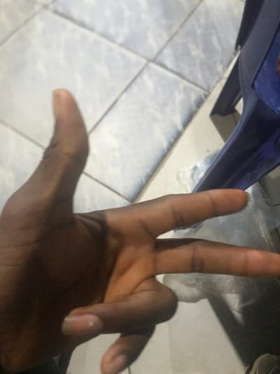 A darkly complected hand appears prominently in the frame. Two fingers are extended upwards, forming a "V" shape. The thumb is stretched outwards, away from the other fingers. The remaining fingers are folded inward. In the background, there is a blue object and a tiled floor with a white pattern. A plastic bag is also visible near the top right.