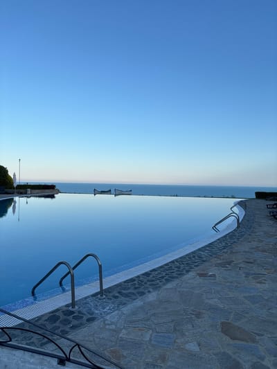 The image shows an infinity pool overlooking the Black Sea in Topola, Bulgaria. The sky is a clear, pale blue, transitioning to a lighter hue near the horizon. Two canoes are visible on the calm, reflective surface of the sea. To the left of the pool, there's a white umbrella, lounge chairs, and lush green bushes. The pool itself is a deep blue, with metal railings and steps leading into the water. The pool deck is paved with irregular, grey flagstones. The overall scene evokes a sense of tranquility and serenity.