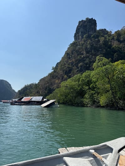 A serene scene features a tranquil body of water reflecting the sky. Lush greenery covers the steep terrain of a rocky cliff face. Several boats with wooden structures are anchored along the shore. A close-up view of a gray boat's interior is on the right, with wooden slats visible. Clear blue sky contrasts with the greenery.