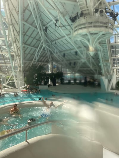 This is an interior shot of the Caldea spa in Les Escaldes, Andorra. The scene focuses on a hot tub area with several people submerged in the turquoise water. Some are swimming or relaxing, while others are closer to the edge of the tub, holding onto a metal railing. The foreground is partially obscured by mist or steam emanating from the water.
The spa's architectural features are prominent, with a high, intricate white structure spanning the ceiling. Lighting and other equipment are suspended from the ceiling. Outside the pool area are trees and the spa's complex's exterior walls. Windows give the space natural light.