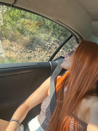 A woman with long, auburn hair is seen from the side, looking out of a car window. She is wearing glasses with thin, silver frames and a leopard-print top. A seatbelt is visible across her chest, and a bracelet adorns her wrist. The background outside the window shows a landscape of trees and rocky terrain, suggesting a rural or natural setting. The lighting indicates it is daytime, with sunlight casting shadows and highlighting the woman's hair. The scene appears to be a snapshot of a journey, possibly a road trip, in Mišići, Montenegro. There is no visible text in the image.