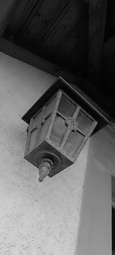 An old, ornate, black metal lantern hangs tilted from a textured white wall, beneath a dark wooden overhang. The lantern has glass panes with decorative metalwork on each side. The wooden structure above consists of planks and beams. A pale, vertical column is visible to the right of the lantern. The scene appears to be outdoors, possibly on the side of a building in Schwaikheim, Germany. There are no people or animals present.