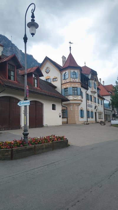 The image depicts the exterior of a building complex in Schwangau, Germany, likely a tourist area given the presence of a "Toiletten" sign. The architecture is distinctive, featuring traditional Bavarian styles with colorful facades, red tile roofs, and charming blue-shuttered windows. In the foreground, a wooden planter box overflows with a vibrant assortment of red, pink, yellow, and white flowers, situated alongside a grey asphalt road. A street lamp with an ornate design stands prominently to the left, casting a shadow. Behind the flowers, two large wooden doors suggest garages or entrances to the buildings. The main building on the right has multiple stories, a turret with a weather vane, and balconies. The background shows a mountainous landscape under a cloudy sky. There are no people visible in the image. The scene is well-maintained and suggests a picturesque, perhaps historical, setting within the town.