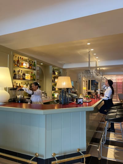 A woman in a white shirt and black apron is working behind a red-topped bar in what appears to be a restaurant or bar. She is standing to the left, using a blender. Another woman in a white shirt and black pants is sitting at the bar to the right, holding a glass and looking up. The bar has a white paneled front and is equipped with bar stools, glassware hanging from the ceiling, and shelves stocked with bottles of alcohol. The background includes more shelves of bottles and decorative items. The scene is well-lit with warm lighting. This establishment is located in Port De Pollença, Spain.