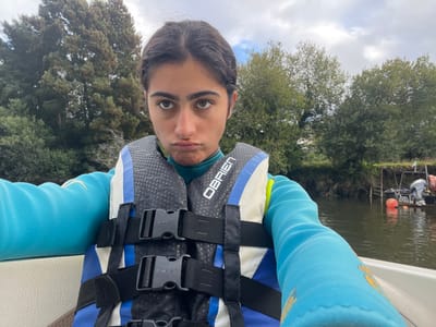 A young individual is depicted in a close-up, selfie-style image, seated in the front of a white boat on a body of water in Vila Nova de Cerveira, Portugal. The individual has dark hair and dark eyes, wearing a teal long-sleeved top and a blue, white, and black O'BRIEN life vest with black straps and buckles. Their lips are pursed in a distinct pout, and they look directly forward at the camera. The sky above is overcast. The background features dense green trees and foliage lining both banks of the calm, dark water. On the right side of the frame, another darker boat with a silver outboard motor is partially visible, moored next to a low wooden structure on the bank. An orange buoy floats in the water near the moored boat, and the name "JOEL" is faintly visible on its stern.