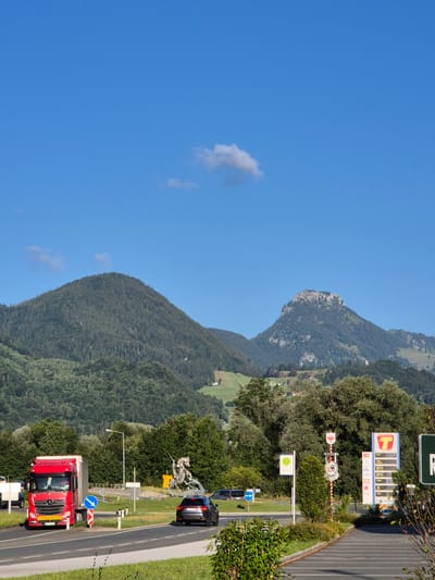 A bright, clear blue sky is visible above a mountainous landscape. The foreground features a road with several vehicles. A large red truck with a white trailer is on the left side of the road, turning. A dark SUV is in the right lane, moving away from the camera. There are traffic signs and a sculpture of a knight on a horse fighting a dragon in the background, situated on a roundabout. A gas station with price displays and signage for "T" fuel is also visible. Trees and green hills form the midground, leading to the imposing, tree-covered mountains in the distance. The overall scene depicts a roadside environment in a mountainous region, under a sunny sky.