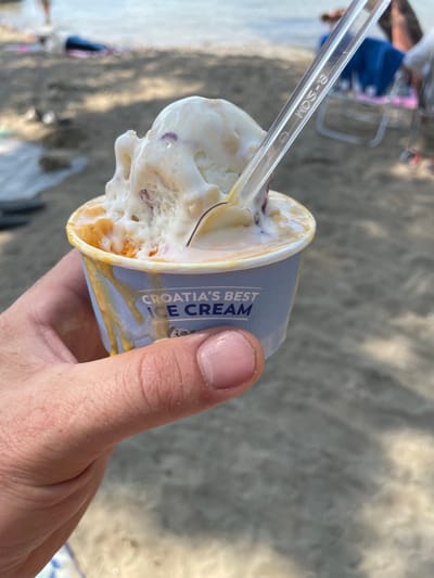 The image shows a close-up of a hand holding a cup of ice cream with a plastic spoon. The ice cream appears to be vanilla-based with chunks of fruit or other additions, and it is melting, with some dripping down the side of the cup. The cup has a blue and white design with text that reads "CROATIA'S BEST ICE CREAM". The background is out of focus, but suggests a beach or outdoor setting with sand and possibly water. The lighting suggests it is daytime, likely sunny, given the bright illumination and shadows. The overall mood seems casual and relaxed, consistent with enjoying a treat outdoors. The spoon has some visible text, "B-SCM", which may be a manufacturer's mark or product code.