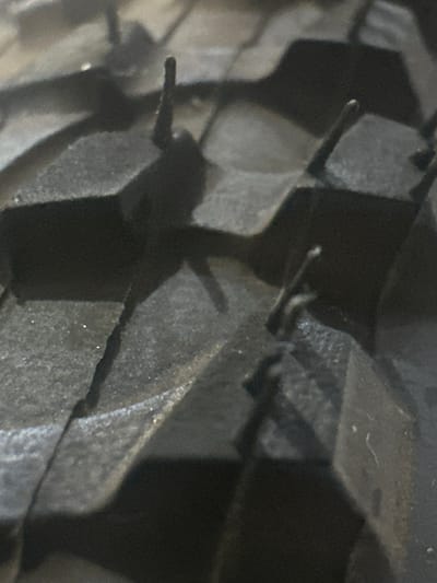 The image shows a close-up, low-angle view of the tread pattern on a tire. The tire appears to be dark in color, likely black rubber, and the tread blocks are deeply grooved and angular, suggesting it is a heavy-duty or off-road tire. Some small, light-colored particles are scattered on the surface of the tire. The lighting is dim, casting shadows that highlight the texture and depth of the tire's design. There are no people or discernible environmental cues to indicate the time of day, weather, or a specific location beyond what can be inferred from the object itself. The image focuses solely on the intricate details of the tire tread. No visible text is present.