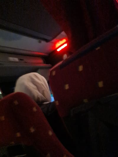 A low-angle shot inside what appears to be a train or bus. Red patterned seats dominate the view. A white baseball cap is visible between the seats. To the left, a dark gray wall features two illuminated red stripes. The overall lighting is dim, creating a claustrophobic feel. The perspective is slightly tilted.