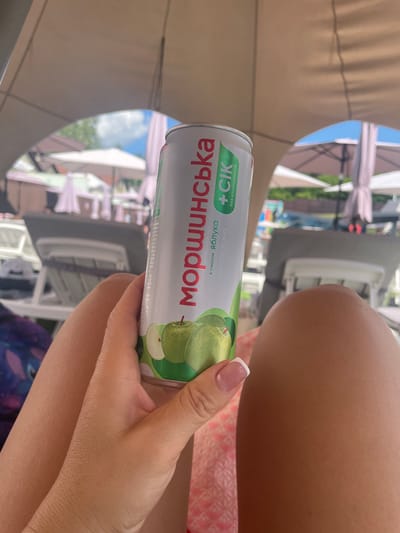 A close-up, eye-level, medium shot captures a person's hand holding a can of "МОРШИНСЬКА + сік" (Morshynska + juice) beverage. The can is predominantly white with red and green lettering. The red text says "МОРШИНСЬКА" vertically along the side. To the right of this text, in green, is "+ сік" and below that, in smaller green text, "без консервантів" (without preservatives). An illustration of sliced green apples is visible on the lower portion of the can. The background shows a blurry outdoor scene, likely a resort or outdoor seating area, with lounge chairs, umbrellas, and some foliage. The person's skin appears tanned, and they have manicured nails with a French tip. The overall setting suggests a relaxed, sunny atmosphere, potentially at a poolside or resort location in or near Zazymia, Ukraine, given the Ukrainian language on the product.