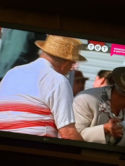 The image shows a close-up of a television screen displaying a scene with several people. In the foreground, a person wearing a straw hat and a white shirt with red stripes is visible from the back and side. To their right, another person is partially visible, wearing a light-colored jacket. A logo with three white circles containing the Cyrillic letters "т", "е", and "т" is displayed on the screen, along with pink text that appears to be in Cyrillic. The background is blurred and appears to be an outdoor setting. The bottom of the television screen shows the "LG" logo. There is no visual information in the image that specifically places the scene in the city of Osolynka, Ukraine.