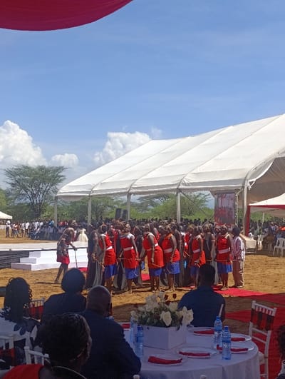 This image depicts an outdoor cultural gathering or ceremony in Marigat, Kenya, characterized by a large white canopy tent structure providing shade.
In the mid-ground, a group of approximately fifteen to twenty individuals, predominantly female, stand in a loose formation on dry, reddish-brown earth. They are dressed in traditional attire, consisting of red tops or shawls, blue skirts or wraps, and multiple beaded necklaces around their necks. One individual, possibly male, similarly dressed in red and blue, stands slightly elevated on a multi-tiered white platform or stage to the left of the main group, holding a long stick or staff. This central group appears to be performing or participating in an organized event.
In the foreground, several guests are seated at white round tables, their backs to the camera. The tables are covered with white tablecloths, set with red placemats, white plates, and bottles of water. A white rectangular planter containing white roses and red flowers serves as a centerpiece on one table. White chairs with red plaid fabric coverings are visible.
In the background, behind the performers and stage, a larger crowd of spectators stands or is seated under additional tent structures. Sparse, dry-looking trees and bushes are visible in the distance under a clear blue sky with scattered white cumulus clouds. A large screen or banner with an image is visible at the back of the primary tent structure. The ground throughout the visible area is dry, exposed soil. Red fabric drapes are visible in the upper left corner, likely part of an overhead decoration.