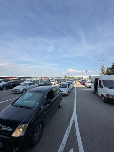 This image presents a wide, slightly elevated perspective of a multi-lane vehicle queue at the Kapitan Andreevo, Bulgaria border crossing, under a partly cloudy, bright blue sky. Numerous passenger vehicles, including sedans, hatchbacks, and vans, are arrayed in multiple parallel lanes, appearing static or slowly advancing. Several large white tour buses are also visible in the background, similarly queued.
In the immediate foreground on the left, a black Opel Meriva minivan is positioned, with its front right headlight illuminated. On the far right, a white commercial van, consistent with a Mercedes-Benz Sprinter model, is visible with its passenger-side sliding door open.
In the mid-ground, a series of low-rise border control buildings with canopy structures are clearly identifiable, positioned across the width of the road. Above and behind these facilities, two prominent flags are displayed: a red flag bearing a white star and crescent (Republic of Turkey) and a blue flag adorned with twelve yellow stars (European Union).
The asphalt road surface exhibits clear white lane markings, indicating organized traffic flow. Small sections of asphalt cracking are visible in the foreground lane. Scattered green trees and tall streetlights are present on the right side of the image, beyond the queued vehicles. People are discernible as occupants within the waiting vehicles; no individuals are observed outside of their cars interacting or moving about freely. The lighting conditions indicate daylight hours.