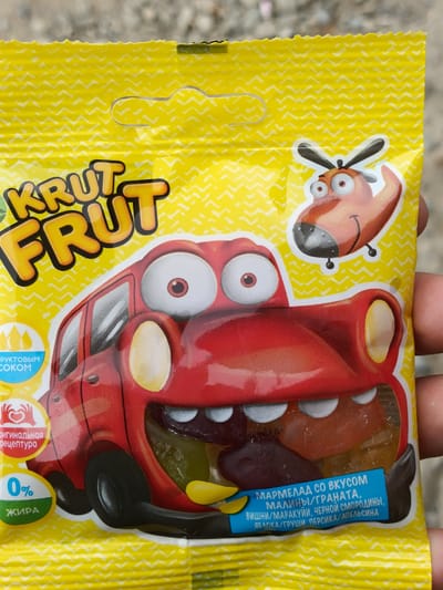 This is a close-up, eye-level shot of a bright yellow package of "KRUT FRUT" fruit jelly candies. The packaging is adorned with a playful, cartoonish design featuring a red car with large, surprised eyes and an anthropomorphic helicopter with googly eyes. The candies themselves are visible inside the packaging, appearing as colorful, translucent jelly pieces.
Prominently displayed on the packaging are several pieces of text in Russian. The brand name "KRUT FRUT" is written in large, bold, yellow and orange lettering. Below this, there are smaller text labels. One circular icon on the left indicates "Фруктовым соком" (with fruit juice) and another below it reads "Оригинальная рецептура" (Original recipe). Further down, a green and white circle highlights "0% ЖИРА" (0% Fat).
A blue rectangular label at the bottom of the image lists the flavors: "МАРМЕЛАД СО ВКУСОМ МАЛИНЫ/ГРАНАТА, ВИШНИ/МАРАКУЙИ, ЧЕРНОЙ СМОРОДИНЫ, ЯБЛОКА/ГРУШИ ПЕРСИКА/АПЕЛЬСИНА" (Marmalade with flavors of raspberry/pomegranate, cherry/passionfruit, blackcurrant, apple/pear, peach/orange).
The packaging has a textured, slightly crinkled surface, and the background behind it is out of focus, suggesting it's being held by a person. The lighting is bright, casting a slight shadow. The overall impression is one of fun and a focus on fruit flavors and healthy attributes. The location context of Razzakov, Kyrgyzstan, doesn't directly influence the visual elements of the product packaging itself, but it provides geographical information about where this product might be found or manufactured.