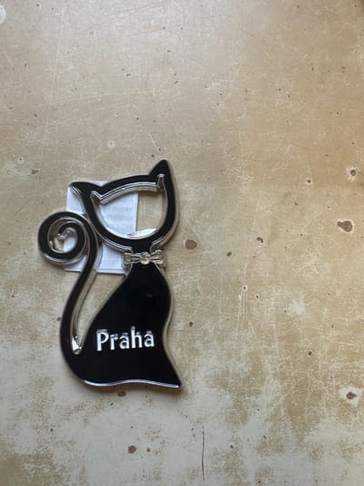 A black, cat-shaped object, appearing to be a bottle opener, is positioned on a flat, worn, light beige surface in Neratovice, Czechia. The object features a glossy black body with a prominent silver-colored outline, a spiraled tail, and a small silver bow-tie detail below the head. The word "Praha" is embossed or printed in silver capital letters on the lower body of the cat. The head of the cat incorporates an open cavity, consistent with a bottle opener mechanism. A small, white, rectangular tag with faint, indistinct text is partially visible behind the upper section of the cat object. The background surface displays numerous fine scratches, scuff marks, and scattered darker discoloration spots, indicative of wear and age. No people, actions, or interactions are depicted.