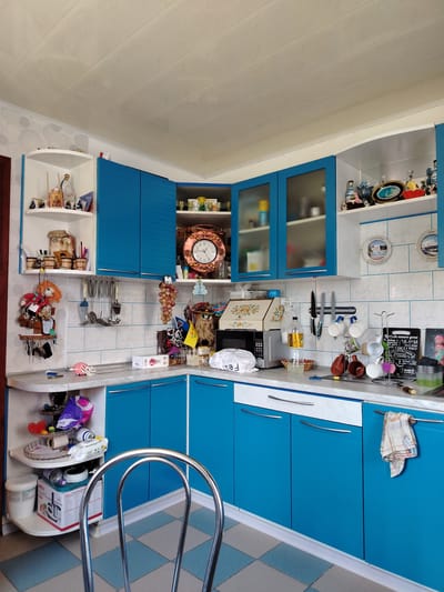 The image shows a brightly lit kitchen with blue cabinets. The kitchen appears to be located in a residential building, possibly an apartment, in Rudzensk, Belarus. The kitchen is well-stocked and organized, with various appliances and utensils visible. The countertops are made of a light-colored material and are lined with several items, including a microwave, a bottle of liquid, and a few containers. The cabinets are a prominent feature of the kitchen, with a mix of closed and open shelves. The walls are tiled with a white, patterned material, and the ceiling is paneled. A dining chair with a chrome frame is positioned in the foreground, suggesting that this is a functional kitchen space. The overall impression is one of a lived-in, practical kitchen environment.