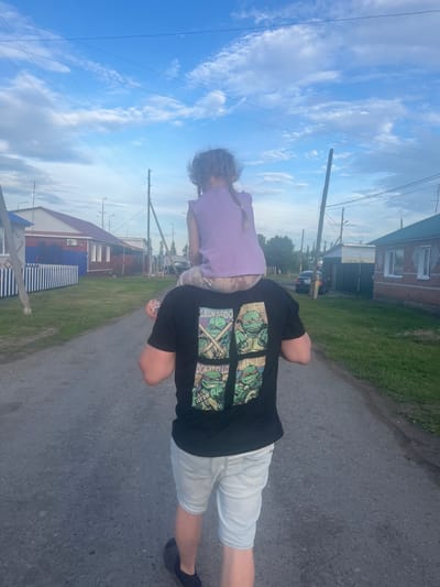 In Mal'tsevo, Russia, an adult from behind carries a young child on their shoulders. The adult wears a black t-shirt with a graphic of four Teenage Mutant Ninja Turtles, and light blue denim shorts. The child is dressed in a light purple t-shirt and patterned leggings. They walk down an asphalt road lined with grassy verges. On the left side of the road are houses with blue and white fences, and on the right, there are more houses and a parked car. The sky above is bright blue with scattered white clouds, and power lines stretch across the frame.