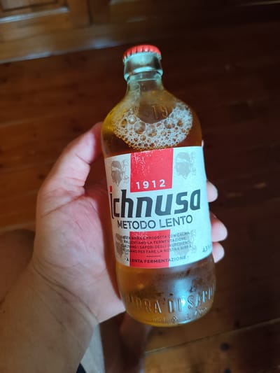 A hand holds a clear glass bottle containing a golden-brown liquid and foam. The bottle has a red cap and a white label with red and black text. The label reads "1912 ICHNUSA METODO LENTO". Below that, it says "QUESTA BIRRA E PRODOTTA CON CALMA, ESALTIAMO LA FERMENTAZIONE. ESALTIAMO I SAPORI DEGLI INGREDIENTI CHE USIAMO PER FARE LA NOSTRA BIRRA A LENTA FERMENTAZIONE.". To the right of this text, "4,3%" is visible. At the bottom of the bottle, embossed text reads "BIRRA DI SARDI" and "50cl". The background is a wooden surface, possibly a table or floor. The image is taken from a slightly low angle, focusing on the bottle and hand. The lighting suggests an indoor environment with warm tones.