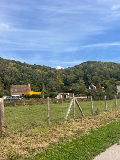 The image depicts a rural-suburban landscape in Vernon, France, on a clear to partly cloudy day. In the foreground, a grassy field is sectioned by a wire fence supported by five weathered wooden posts, with a prominent A-frame support structure in the center-right of the fence line. The grass in the immediate foreground is a mix of green and dry, matted brown patches, bordered on the far right by a strip of greener turf alongside an unpaved or light-colored pathway.
Beyond the field, the midground features two residential structures against a backdrop of heavily wooded hills. To the left, a two-story house with a reddish-brown tiled roof and light-colored walls is visible, with a dark grey SUV-style vehicle parked in front. Adjacent to this house, a distinctive bright yellow rectangular structure stands out. Further right, a second house, smaller and partially obscured by trees, has a lighter-colored roof and walls. A circular red "30" speed limit sign is visible on the far right.
The background is dominated by a large, undulating hill covered with dense deciduous forest, displaying a mix of green, yellow, and subtle orange foliage, indicative of early autumn. The sky above is a light blue with scattered, wispy white clouds. No individuals or discernible actions are present in the scene.