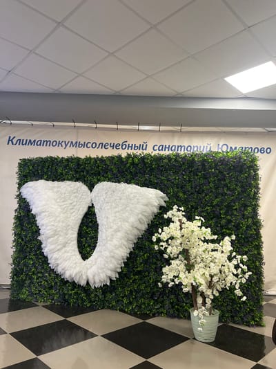 The image displays an indoor decorative scene, likely within a public or commercial building. The central feature is a wall adorned with artificial green foliage, resembling a hedge. Mounted on this green wall are a pair of large, white, feathery angel wings. To the right, in front of the green wall, sits a light blue, vertically striped vase holding a plant with abundant white blossoms, possibly artificial cherry or apple blossoms.
Above the decorative wall, a white banner runs vertically along the left side. This banner features blue Russian text that reads: "Климатокумысолечебный санаторий Юматово," which translates to "Climate and Kumys Therapy Sanatorium Yumatovo." The banner is secured with black ties.
The ceiling above is a light-colored, grid-patterned acoustic tile ceiling, featuring a large, bright rectangular light fixture that illuminates the area. The floor visible in the foreground and to the right has a distinctive black and light grey/white checkered pattern. No people or specific activities are present, suggesting a static decorative installation. The bright lighting indicates an indoor setting during operational hours. The location cues strongly point to a sanatorium named Yumatovo, specializing in climate and kumys therapy.