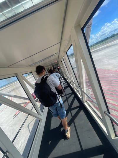 The image shows a group of people walking through an airport jet bridge towards an airplane. The perspective is from behind one of the travelers, a male with a black backpack and denim shorts, facing forward. The jet bridge has large glass windows on the right side, offering a view of the tarmac, runway, and a cloudy blue sky. Red lines are painted on the tarmac. The environment appears to be an airport located in Mai Khao, Thailand, suggested by the tropical landscape visible in the distance. The people are queuing and moving in a single file line, implying they are boarding the aircraft.
