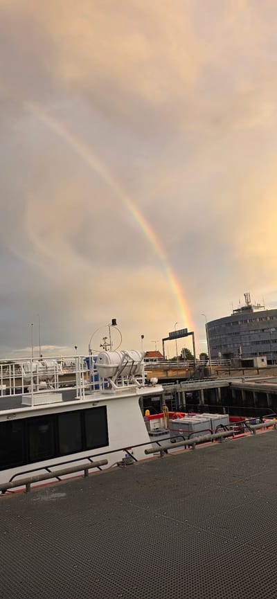 A vertical, eye-level, outdoor photograph captures a double rainbow arching across a muted orange and gray sky. The rainbow appears in the upper portion of the frame, originating from the left and extending towards the right, with a secondary, fainter rainbow visible above it. In the mid-ground, a white ferryboat with life rafts is docked. Its deck is visible, featuring railing and a dark-colored surface. Beyond the ferry, a bridge spans over a body of water. To the right of the bridge, a large, round, gray building with multiple windows and an antenna array on its roof is situated. The scene appears to be a harbor or port in Den Helder, Netherlands, given the presence of ferry operations and maritime infrastructure. The lighting suggests either dawn or dusk.
