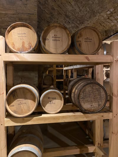 The image depicts a barrel aging cellar in Valmiermuiža, Latvia, characterized by rustic stone and brick arched ceilings. Numerous wooden barrels are systematically stored on multi-tiered, light-colored wooden shelving units.

On the top shelf, from left to right, three barrels are positioned. The leftmost barrel, labeled "№ 25," displays markings "03072024" and "10052018" with a dark stain at its lower front. The adjacent barrel, "№ 26," is marked "23052018," "03072024," and "GRAMATS 03.09.2018." The third barrel, "№ 27," features identical markings: "23052018," "03072024," and "GRAMATS 03.09.2018."

The middle shelf showcases three more barrels. The leftmost barrel, "№ 31," is inscribed with "22062018," "28082025," and "GRAMATS 05.09.2018." Next to it, barrel "№ 32" bears the dates "01112018" and "28082025." The rightmost barrel on this shelf, "№ 33," is darker and extensively hand-marked with details including "Double Barrel," "Texas Bourbon Master," "# 5725," "1st Barrel Mc. B. M. 30 gal Filled @ 114 oz Sept 2 2013," "2nd Barrel Intra Kelvin 27 gal Filled on Oct 6 2015," and "Distilled Ref," along with the date "14.08.2018" and "08."

A partially visible barrel lies horizontally on the bottom shelf. In the background, additional barrels are stacked on similar wooden shelves, and dark bottles are also visible, indicating further storage. Two individuals are partially obscured behind the shelving unit in the mid-ground; one figure on the left appears to be looking right, while another on the right, seemingly wearing a headset, looks left. A third, less distinct figure is visible between them. The overall lighting is dim, suggesting a controlled environment for aging.